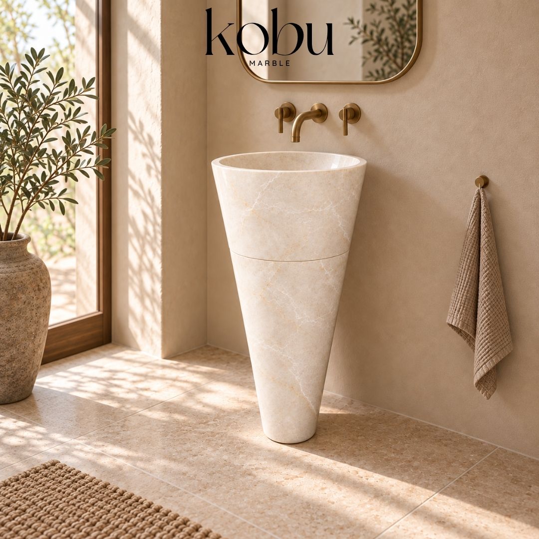 Beige marble pedestal sink with a wall-mounted brass faucet in a sunlit bathroom.